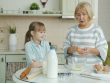 Senior woman is cooking pastry in kitchen teaching little girl discussing ingredients and breaking eggs in bowl. Cookery, family and people concept.