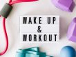 Top view closeup composition of wake up and workout inscription on light box surface among expanders and dumbbells and massage ball on white background